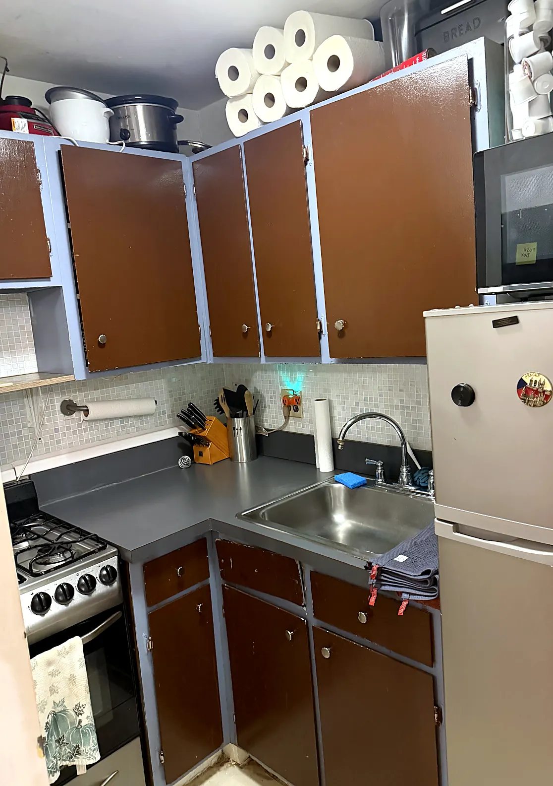 Kitchen in June 2026: brown cabinet doors, grey laminate counter, mosaic backsplash, paper towels stacked on top